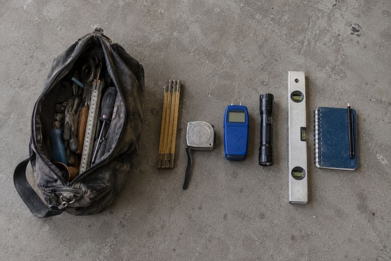 Werkzeugtasche mit Zollstock, Maßband, Feuchtemessgerät, Taschenlampe, Wasserwaage und Notizbuch auf einem Betonboden