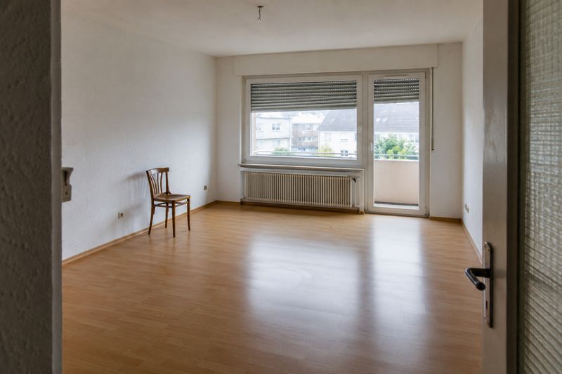 Leeres Wohnzimmer einer Eigentumswohnung mit Laminatboden, Heizkörper unter dem Fenster und Balkontür